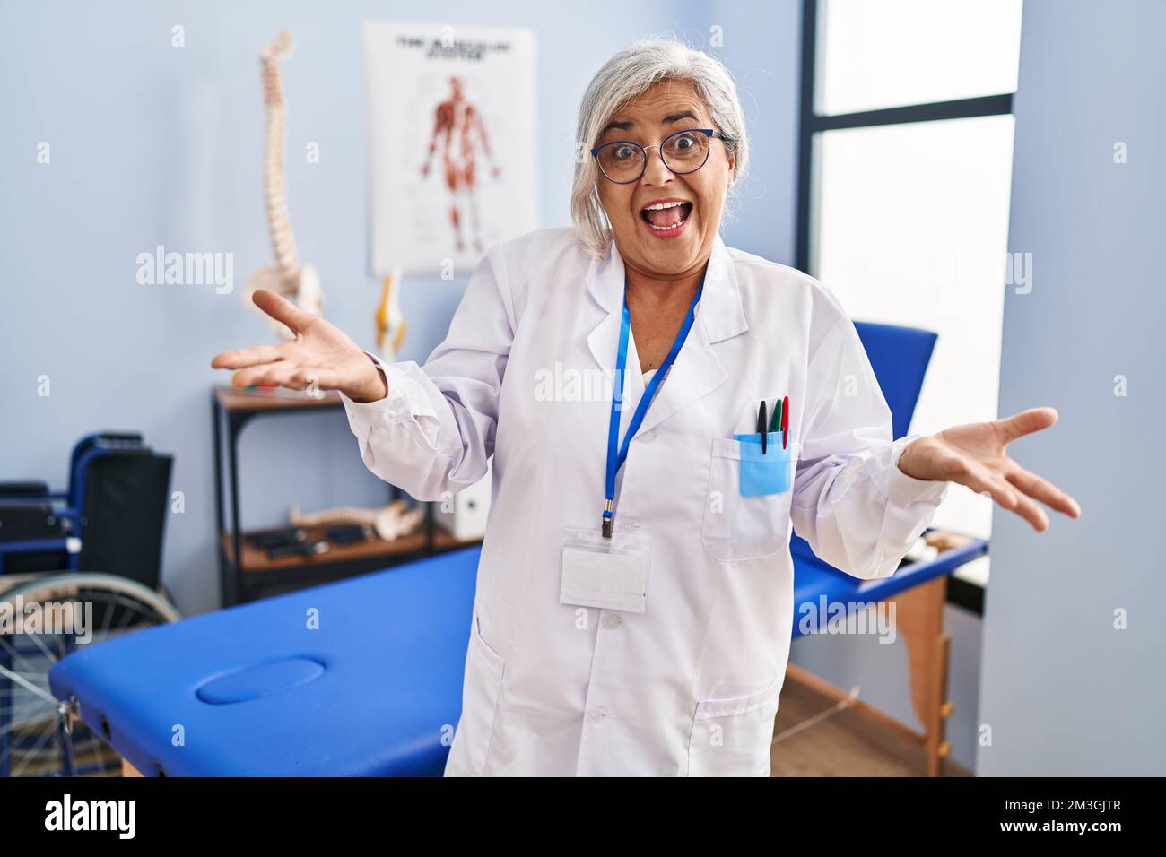 Femme d'âge moyen avec cheveux gris travaillant à la clinique de récupération de douleur souriant gai avec les bras ouverts comme accueil amical, positif et confiant salutations Banque D'Images