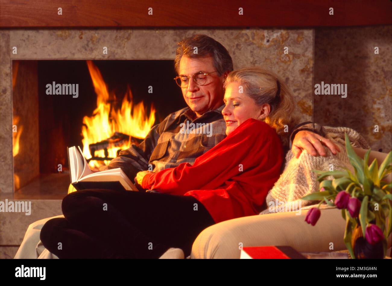 Un couple plus âgé s'est emmêlé sur le canapé en lisant un livre devant un feu de bois dans la cheminée Banque D'Images