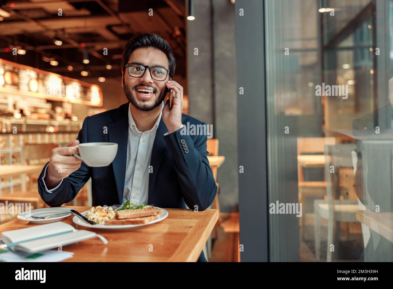 Beau homme d'affaires indien en lunettes parle téléphone avec le client pendant le déjeuner dans un café confortable Banque D'Images