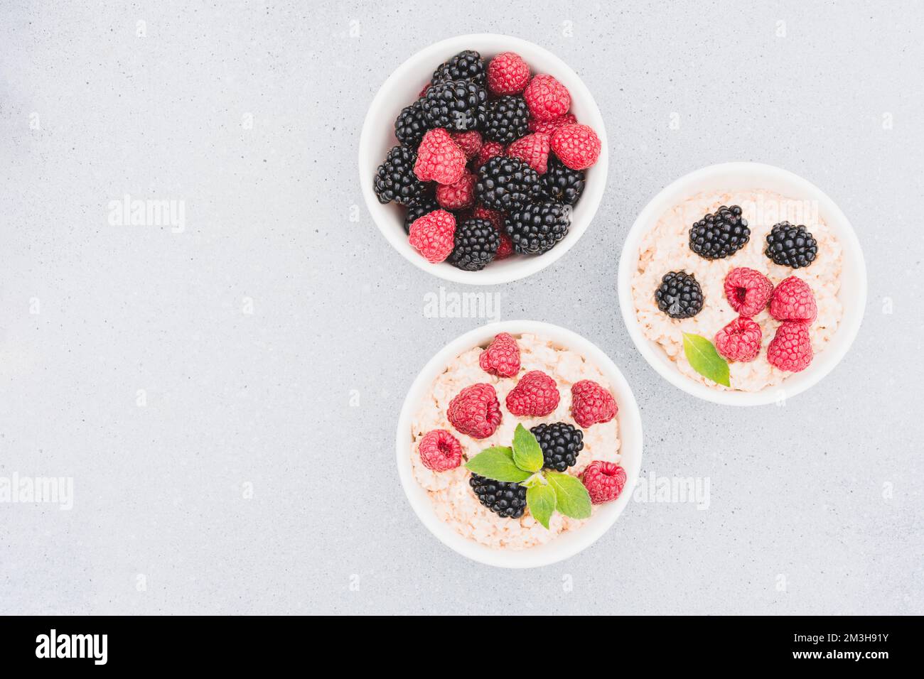 Porridge de flocons d'avoine garni de diverses baies dans des bols blancs sur fond de table en béton avec espace de copie. Recette de porridge du matin Banque D'Images