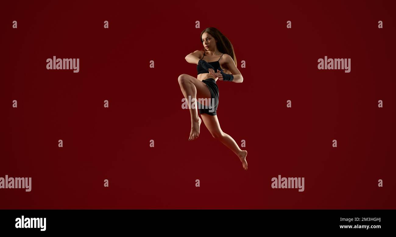 Vue latérale d'une femme sportive brunette avec des cheveux longs sautant haut, sur fond rouge studio. Assez forte d'entraînement de femme dans l'air. Concept des arts martiaux Banque D'Images