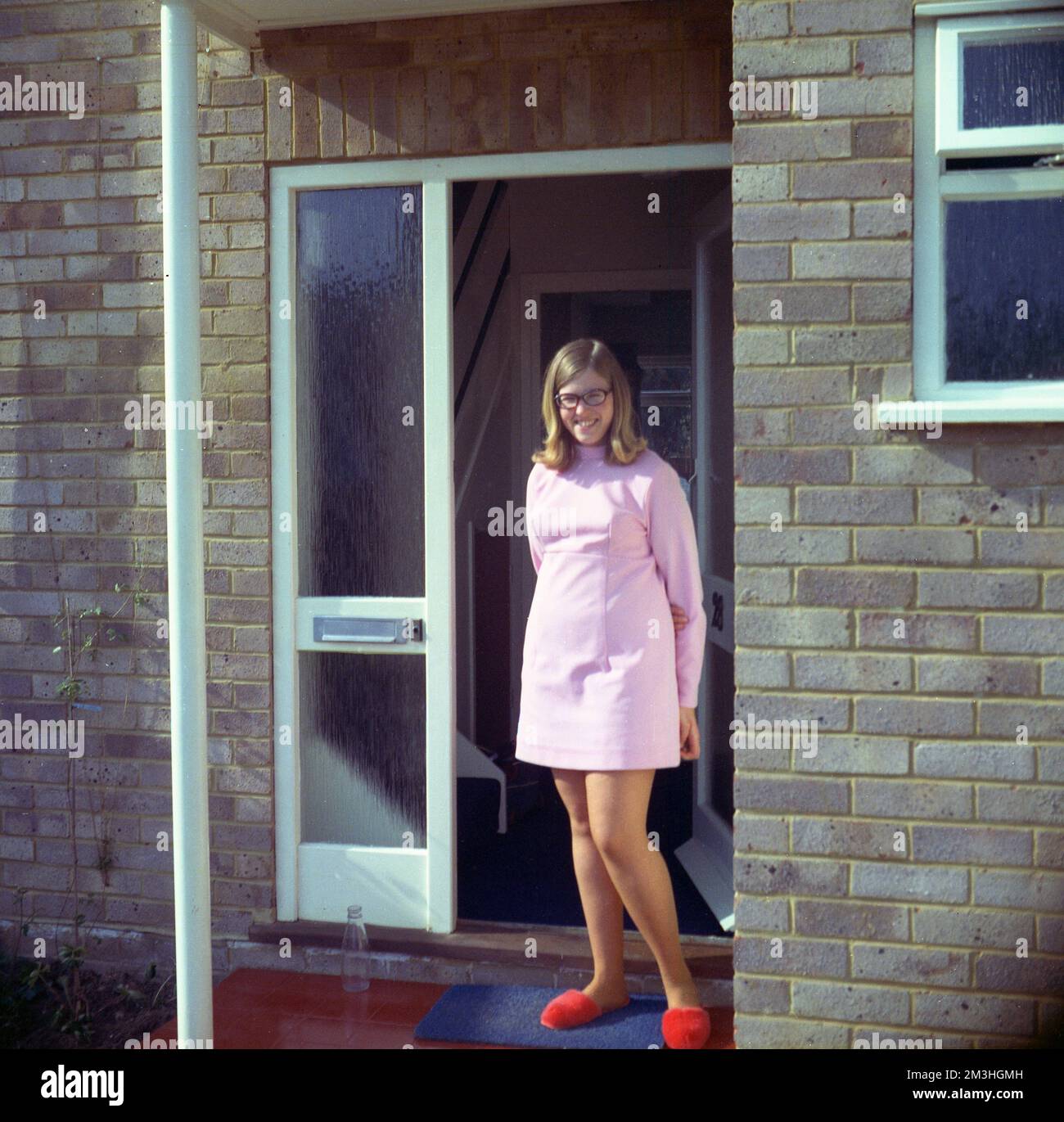 1970, historique, une jeune femme dans sa vingtaine debout pour une photo à l'entrée d'une maison, portant une robe courte rose, à la mode dans cette époque et sur ses pieds, rouge duveteux pantoufles, Angleterre, Royaume-Uni, une bouteille de lait vide est à la porte Banque D'Images