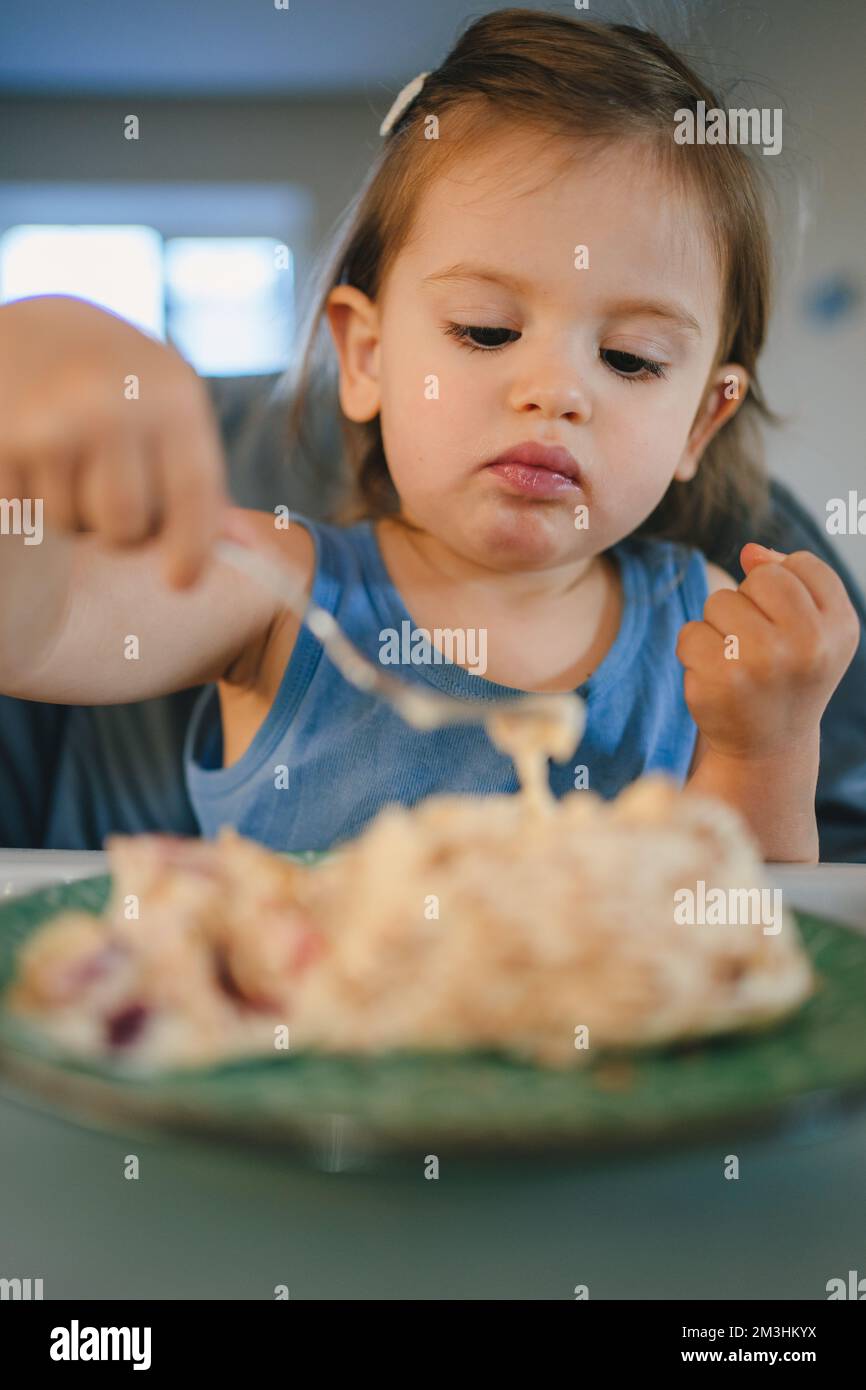 Petite fille de bébé caucasien mangeant de la nourriture solide avec fourche assis sur une chaise haute. Une alimentation saine. Bonne famille. Banque D'Images