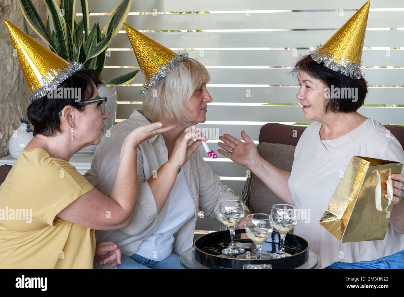 Des femmes âgées heureuses portant des chapeaux de fête buvant du vin, riant, échangeant des cadeaux de Noël, célébrant ensemble dans le patio arrière-cour. Femme d'âge moyen avec chapeau d'anniversaire envoi baiser soufflant. Banque D'Images
