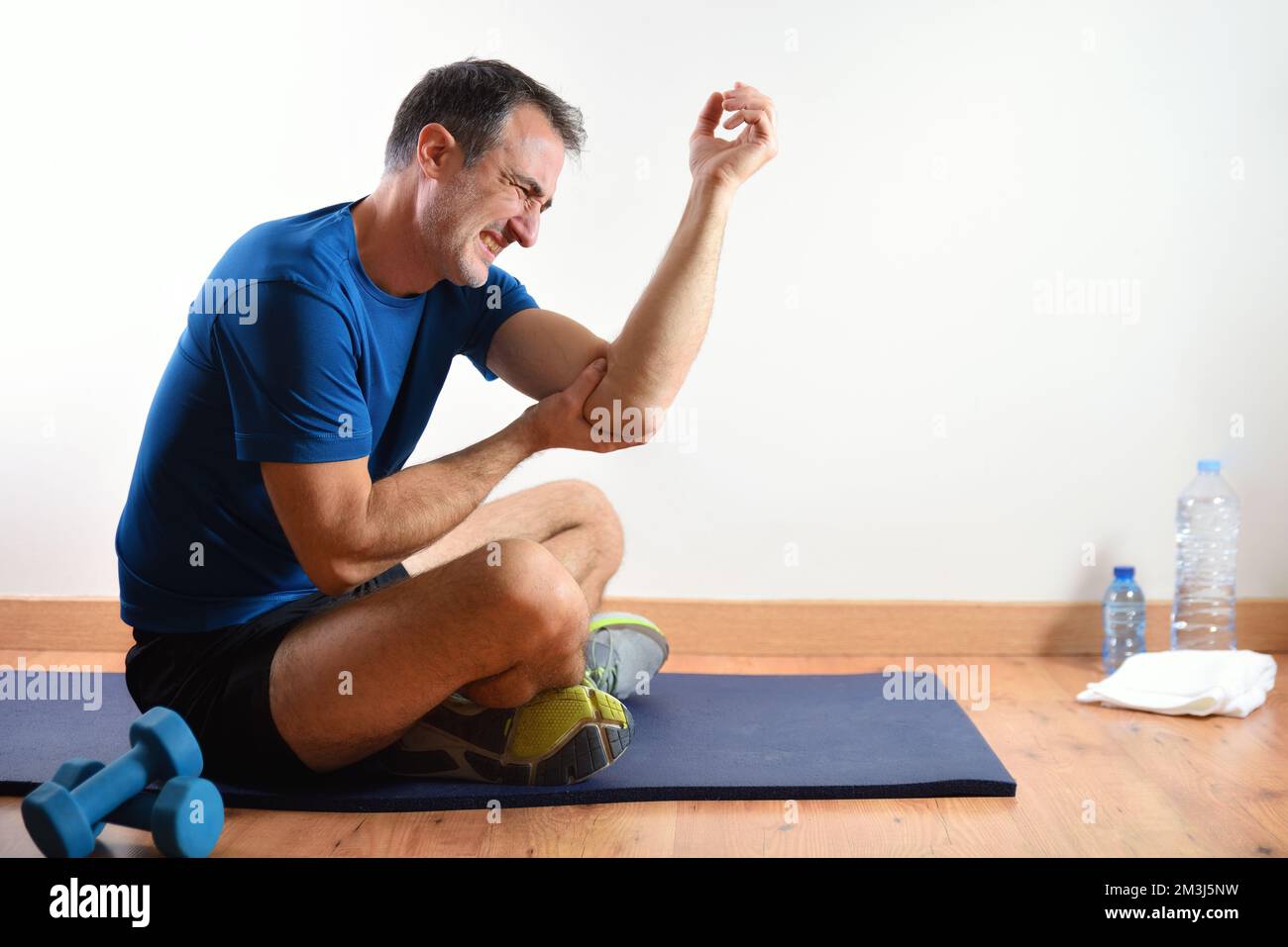 Homme faisant du sport à l'intérieur se plaignant de douleurs au coude assis sur un tapis. Vue latérale Banque D'Images