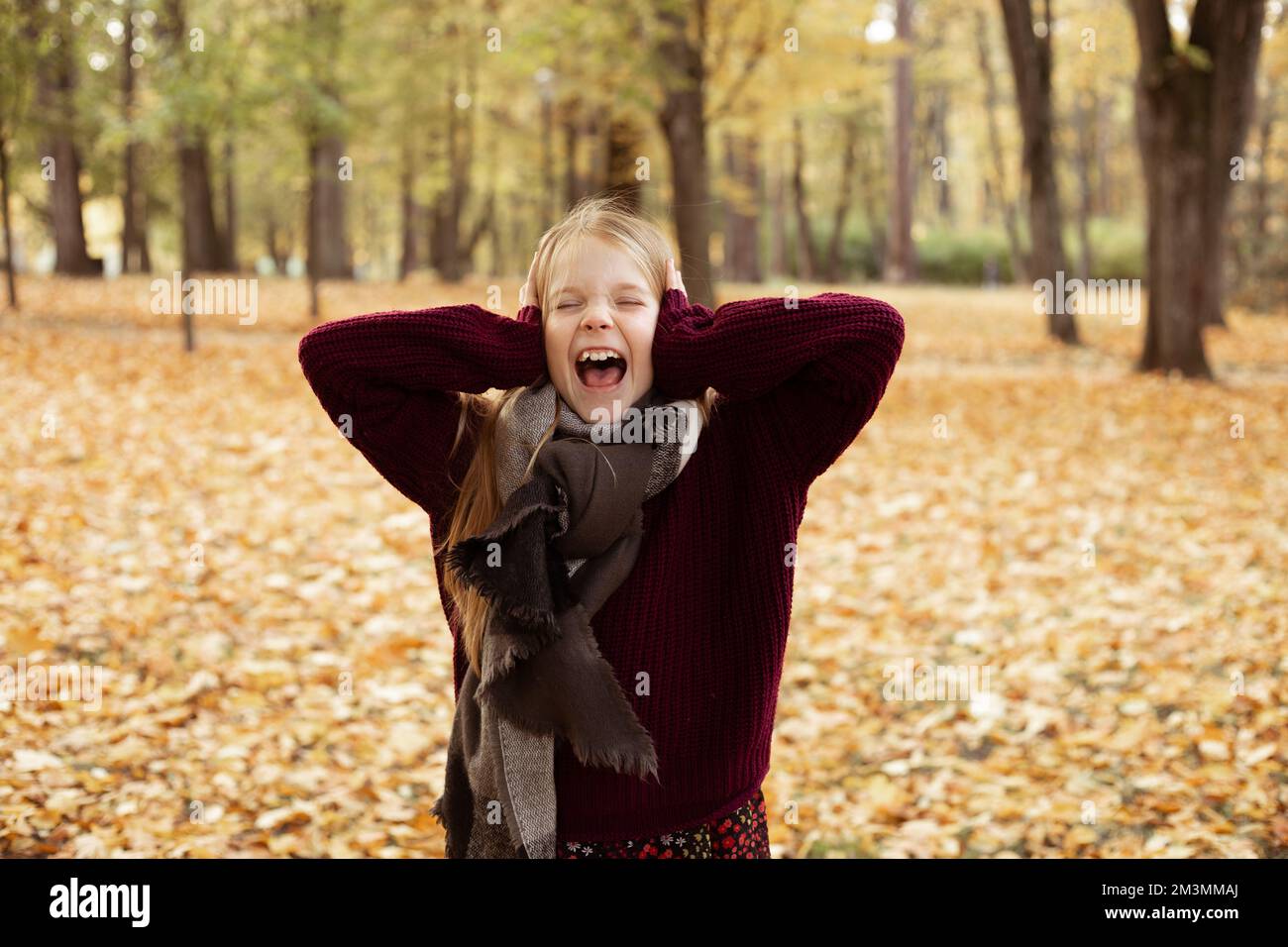 Portrait d'une jeune fille folle debout au milieu de feuilles d'érable jaune dans le parc, hurlant, couvrant les oreilles avec les mains. Banque D'Images