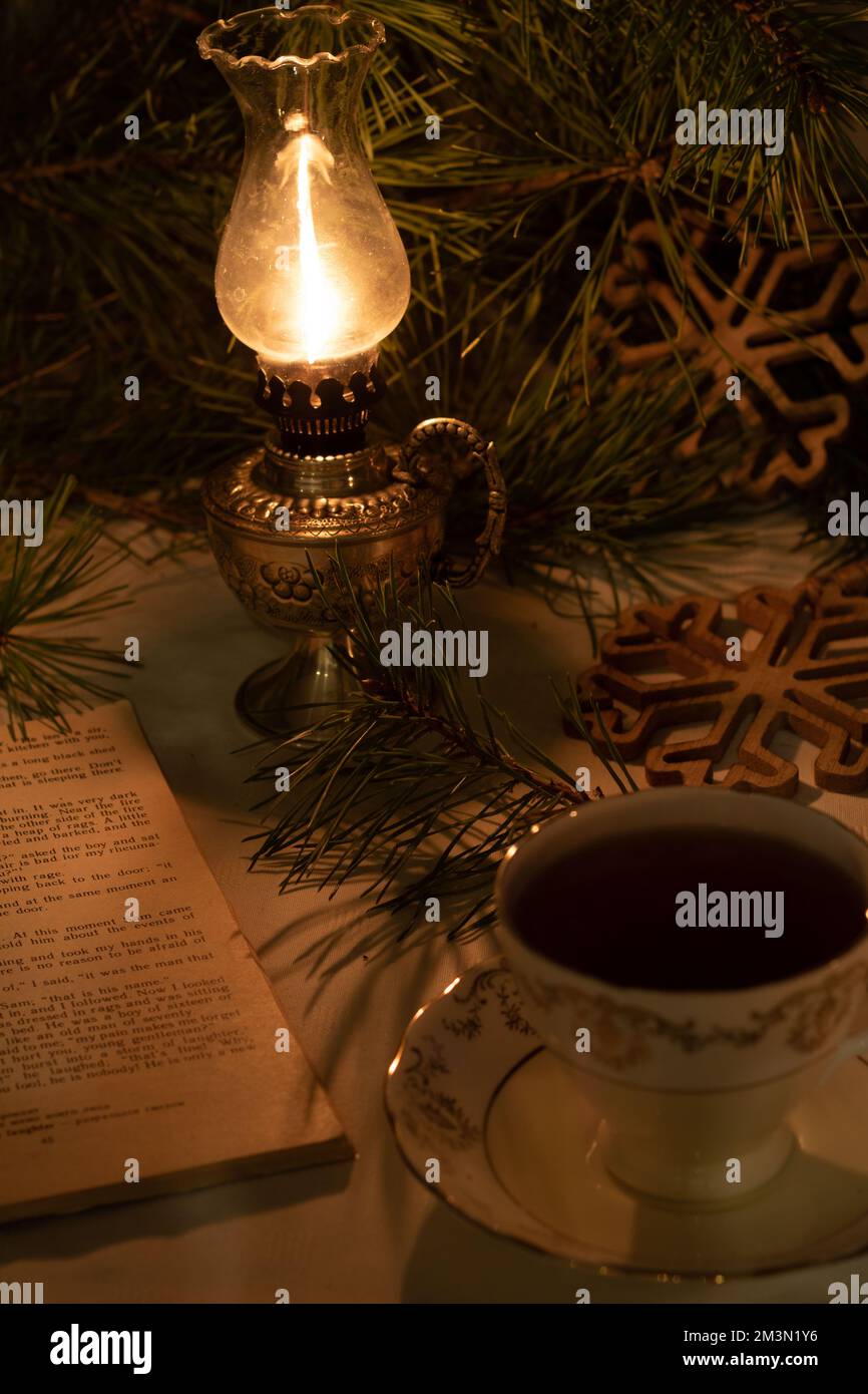 photo sur la table est une lampe à huile brûlante à côté d'une tasse de thé rétro et un vieux livre ouvert dans le fond des flocons de neige en bois sur une branche d'épinette Banque D'Images