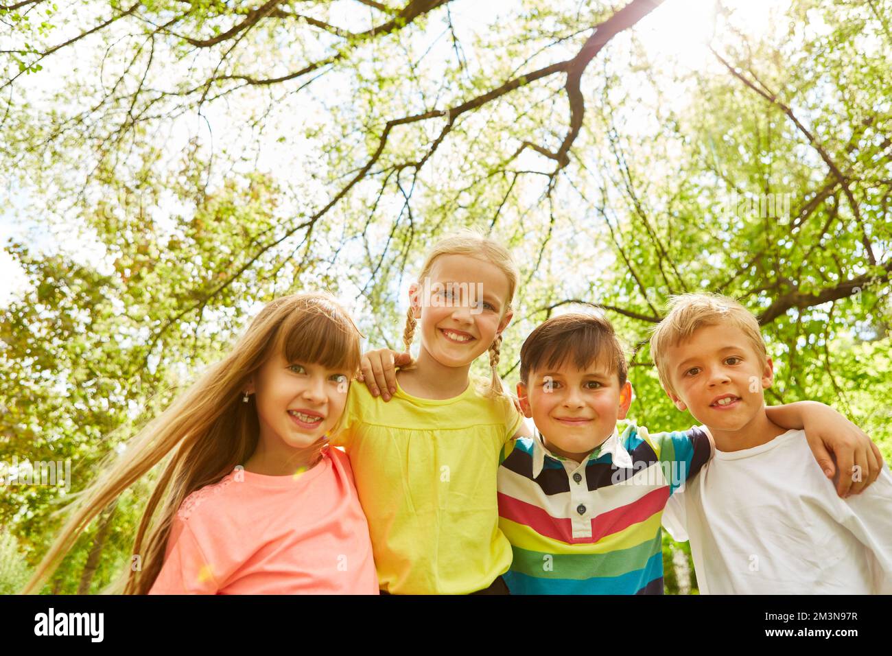 Portrait de garçons et de filles souriants avec les bras autour debout devant les arbres dans le jardin Banque D'Images Portrait de garçons et de filles souriants avec les bras autour debout devant les arbres dans le jardin Banque D'Images