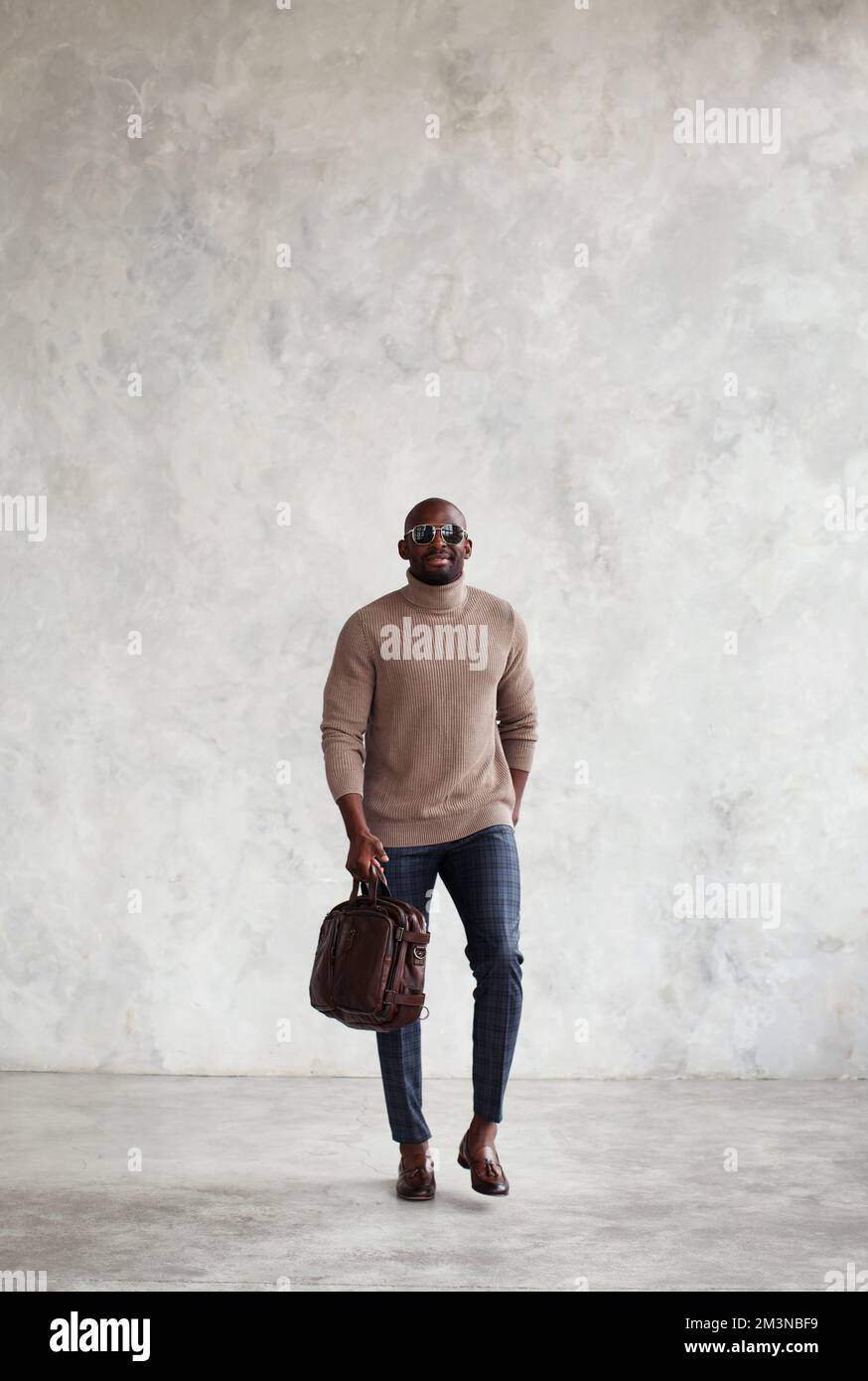 Homme long tendance marche contre un mur gris, porte des vêtements élégants, un sac en cuir marron et des mocassins, un pull à col roulé. Hommes style de mode Banque D'Images