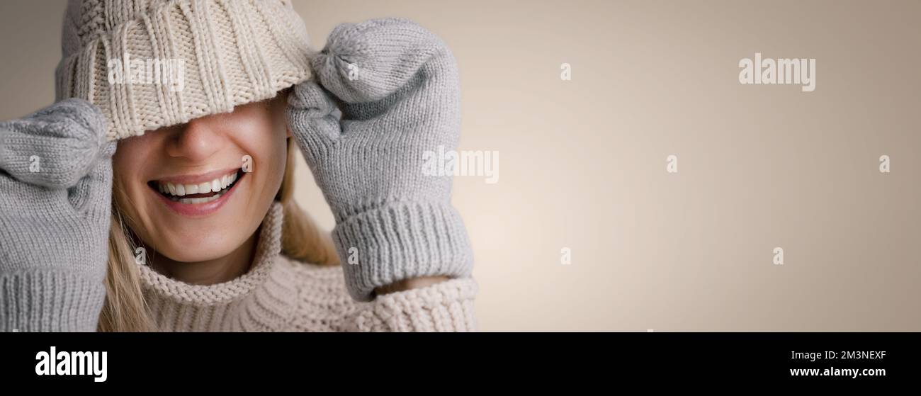 femme souriante portant des vêtements chauds se cachant face sous un bonnet en laine tricoté sur fond beige. vêtements d'hiver tendance. bannière avec espace de copie Banque D'Images