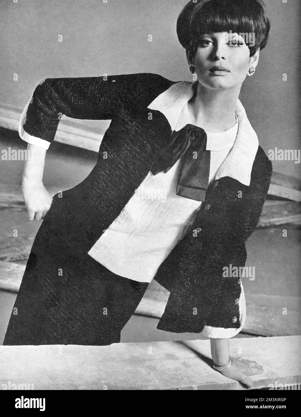 Un costume en chenille noire sucette, la veste avec trois boutons hauts et une jupe portefeuille. Le chemisier est en satin blanc nervuré avec un noeud en satin noir floppy et les poignets de la veste sont fendus haut. Les boucles d'oreilles centrées en jade entourées de perles sont de Bijoux Christian Dior at Woollands et le bracelet plaqué or de Creations grosse at Marshall &amp; Snelgrove. Date: 1965 Banque D'Images