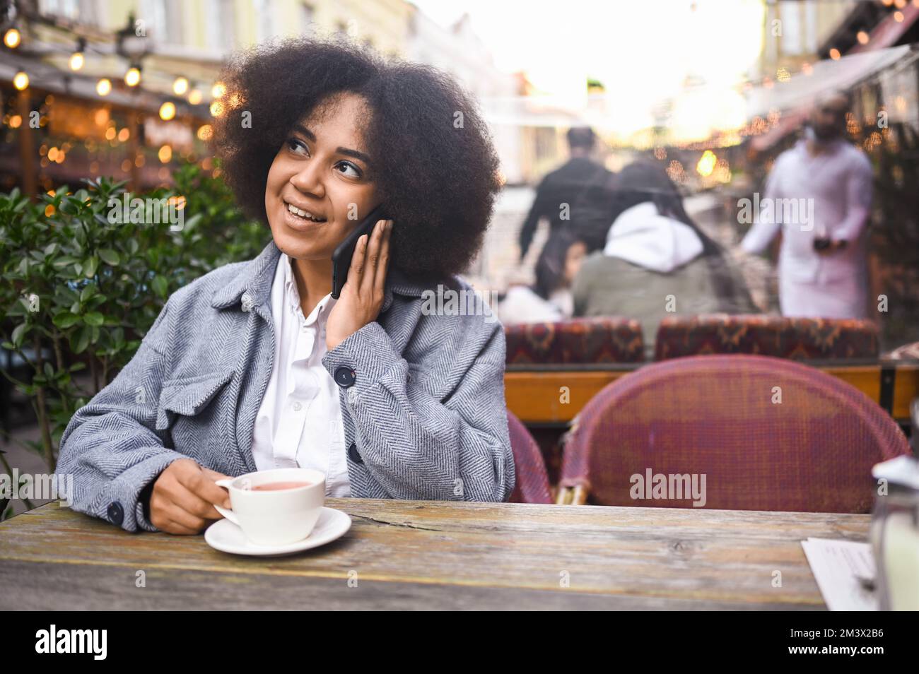 Mode Street style portrait de jeune beauté naturelle attrayante Afro-américaine femme avec des cheveux afro dans la veste tweed parle téléphone à l'extérieur dans le café-terrasse.Bonne dame avec une tasse de café dans la grande ville Banque D'Images