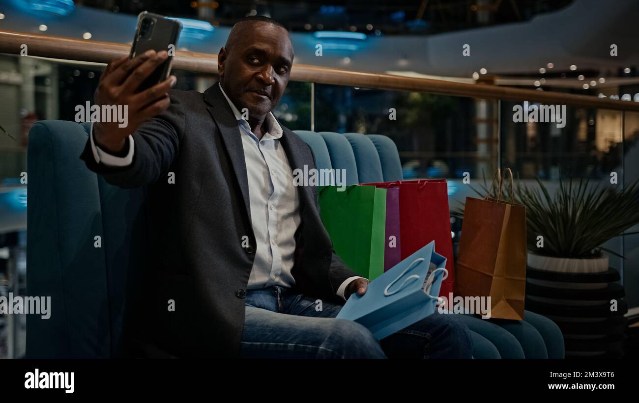 Homme d'âge moyen homme africain acheteur client blogueur influenceur vlogger avec des sacs d'achats assis dans le centre commercial appel vidéo faire de la photographie avec Banque D'Images