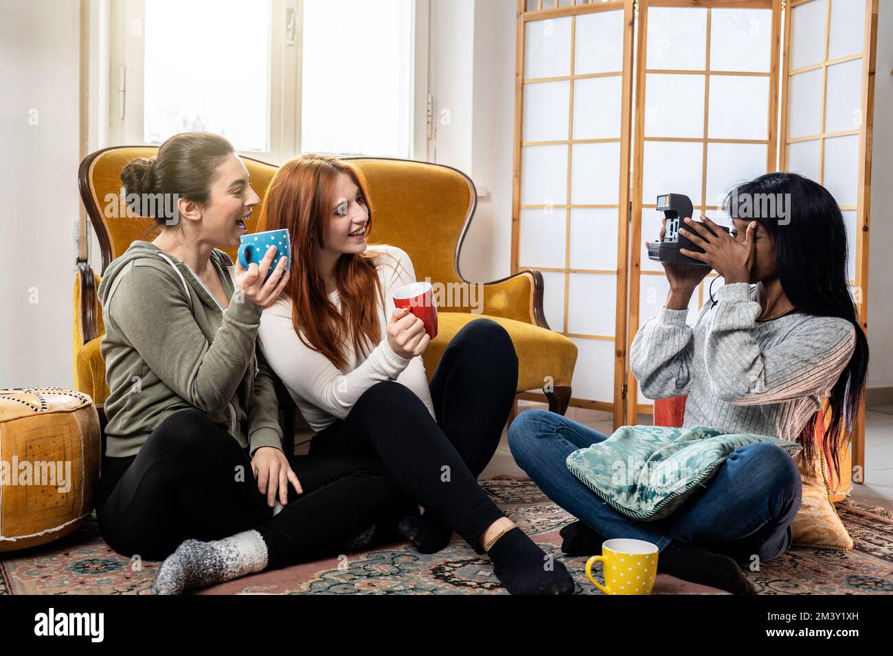 Des amies multiculturelles et gaies qui profitent de leur temps libre à la maison - des jeunes femmes heureuses qui boivent du café ou du thé et qui prennent des photos avec vint Banque D'Images