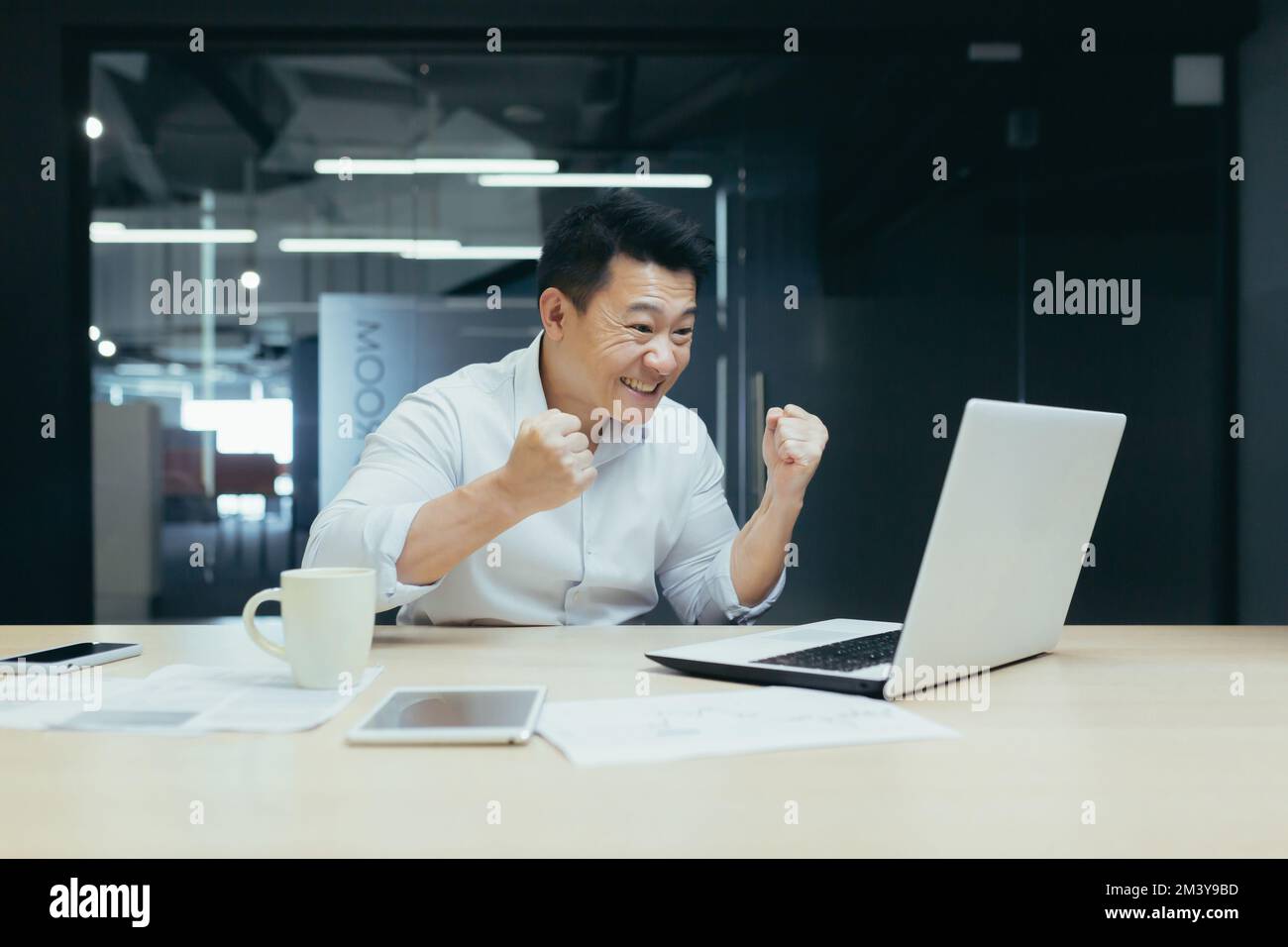 Un jeune asiatique est assis au bureau à la table, pendant les heures de travail il regarde un match de football, des compétitions sportives, fait des Paris, des acclamations et des réjouissances sur son ordinateur portable. Banque D'Images