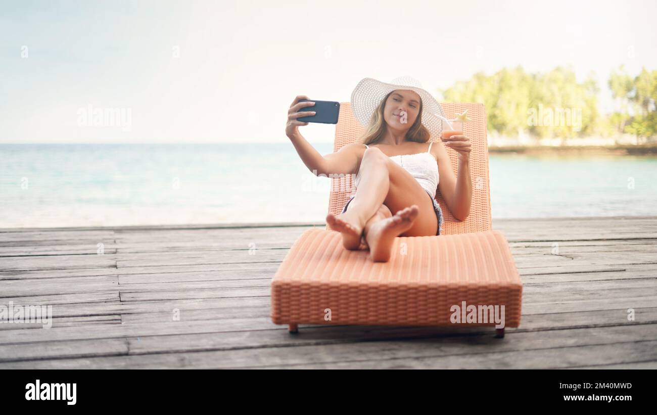 Je ne peux pas voyager et ne pas prendre de selfies. Prise de vue en longueur d'une jeune femme attrayante assise sur un transat et prenant des selfies tout en tenant un Banque D'Images