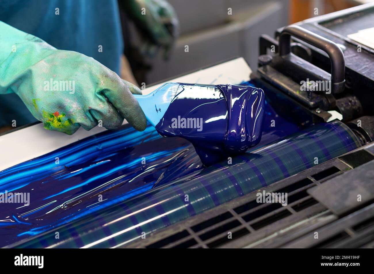 Par dessus l'employé anonyme dans des gants en caoutchouc sale étalant l'encre bleue sur les rouleaux de l'imprimante offset industrielle pendant le travail dans l'atelier de typographie Banque D'Images