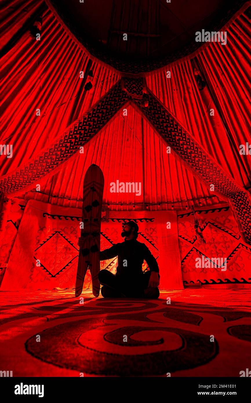 Silhouette de l'homme tenant avec le snowboard séparé assis avec des jambes croisées à l'intérieur de la maison nomade de Yurt contre la moquette avec des motifs ethniques illuminés par la lumière rouge Banque D'Images