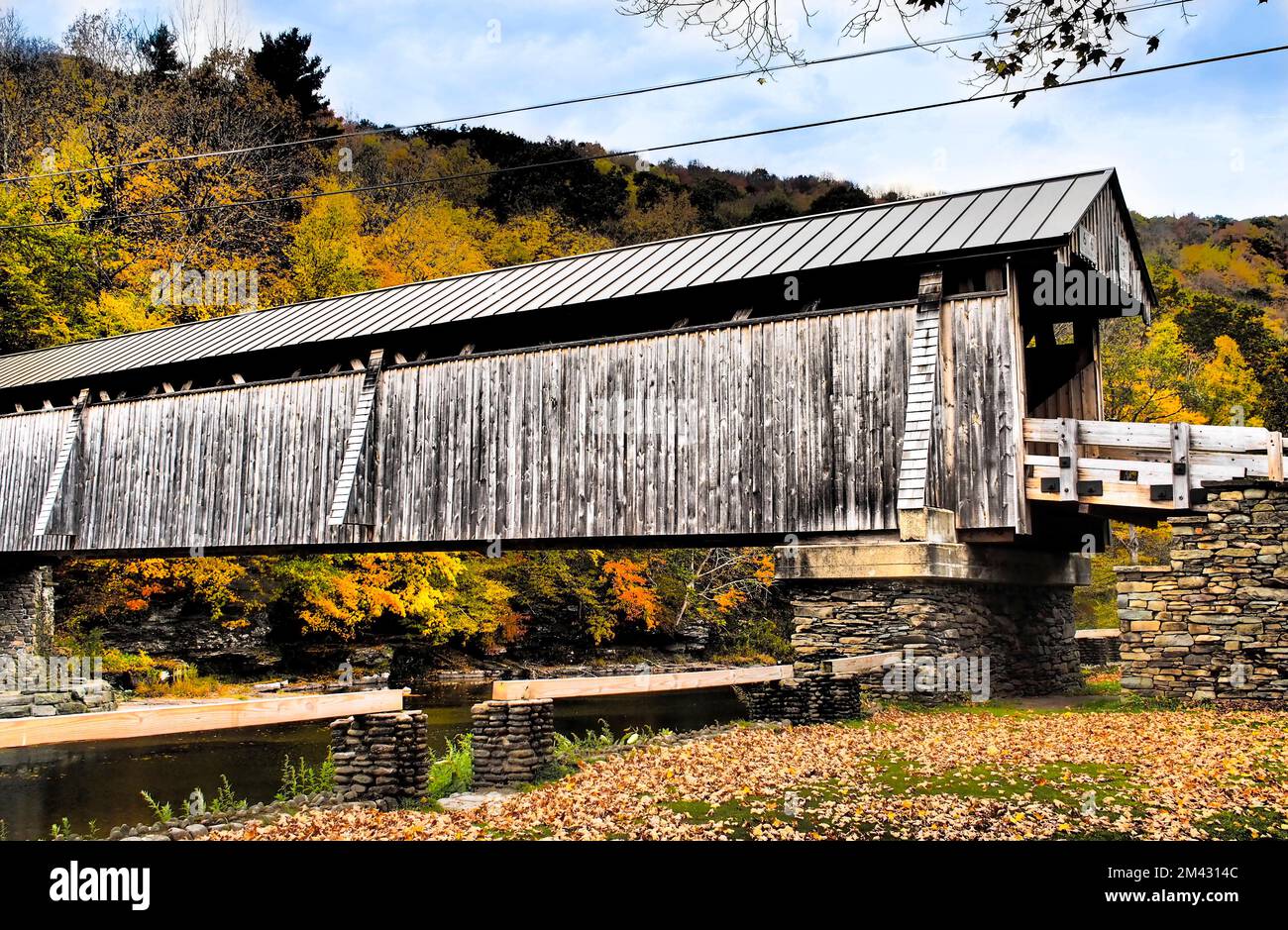 Pont couvert rural au-dessus d'une rivière calme, couleurs vives des feuilles d'automne dans les collines boisées et ciel bleu. Avec filtres pour un effet carte postale rétro. Banque D'Images