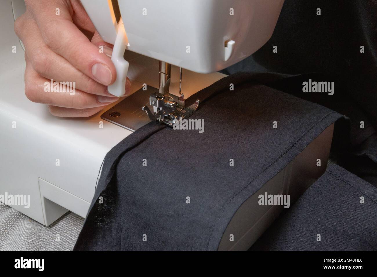 La couturière cousue des vêtements en tissu noir sur une machine à coudre. Lumière de la lampe matérielle intégrée. Aiguille en acier avec boucle et pied de pression. fermer Banque D'Images