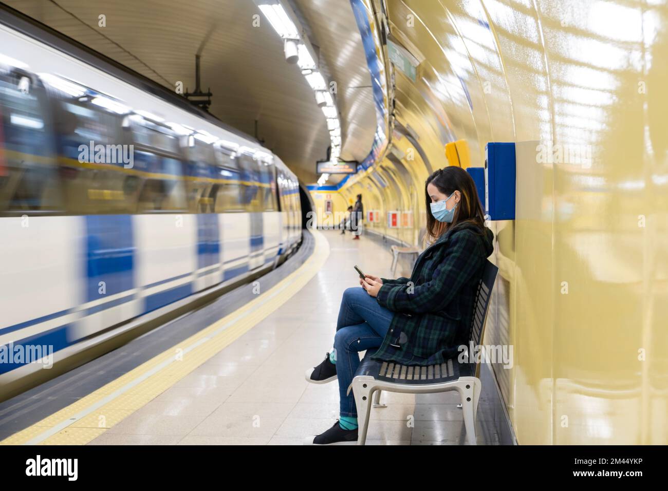 Jeune femme latine assise et portant un masque chirurgical avec un smartphone en attendant dans le métro ou la gare Banque D'Images