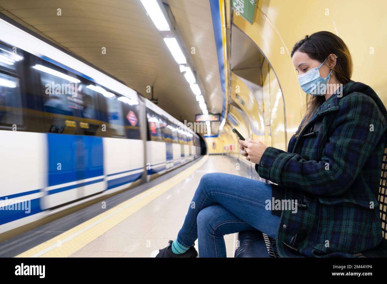 Jeune femme latine assise et portant un masque chirurgical avec un smartphone en attendant dans le métro ou la gare Banque D'Images