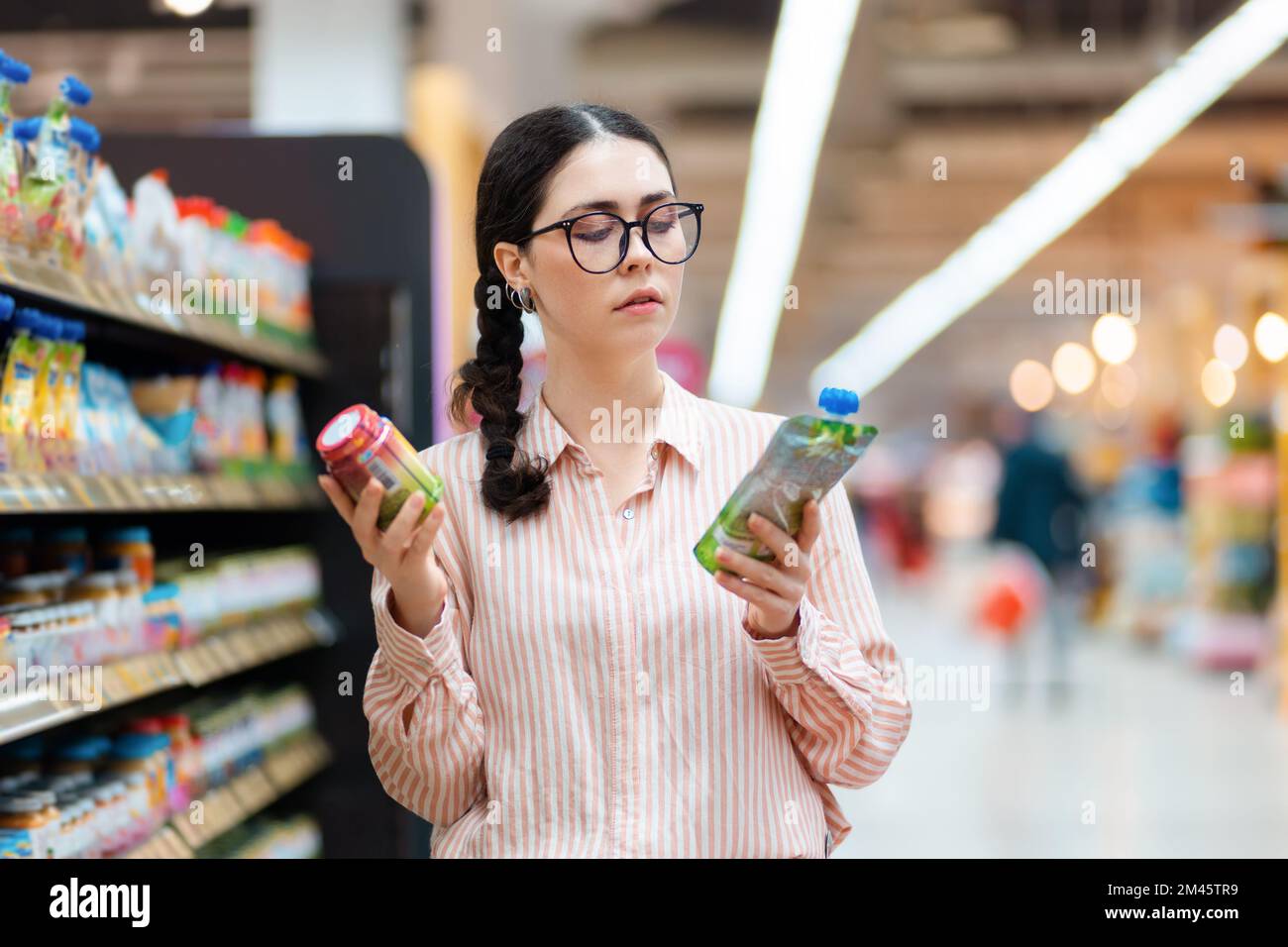 Portrait d'une jeune femme caucasienne portant des lunettes de vue tient les produits dans les mains et ne peut pas faire le choix. Concept de shopping dans les supermarchés et le consumérisme. Banque D'Images