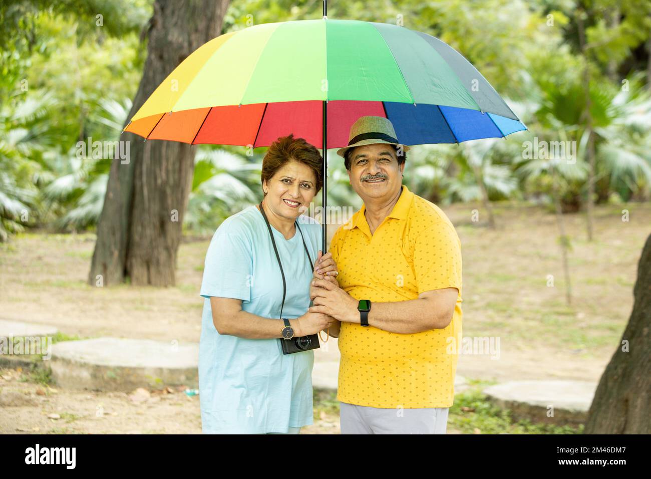 Joyeux couple indien senior debout sous un grand parapluie coloré portant des habits décontractés et un chapeau à l'extérieur du parc, vieux retraités appréciant des vacances. Banque D'Images
