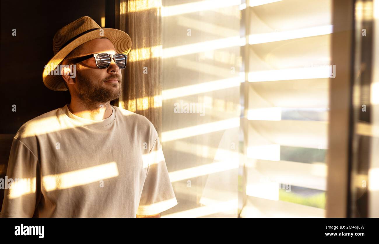 Vêtements décontractés homme avec une barbe courte portant un chapeau de paille et des lunettes de soleil debout dans la chambre et regardant à travers les stores de fenêtre. Banque D'Images
