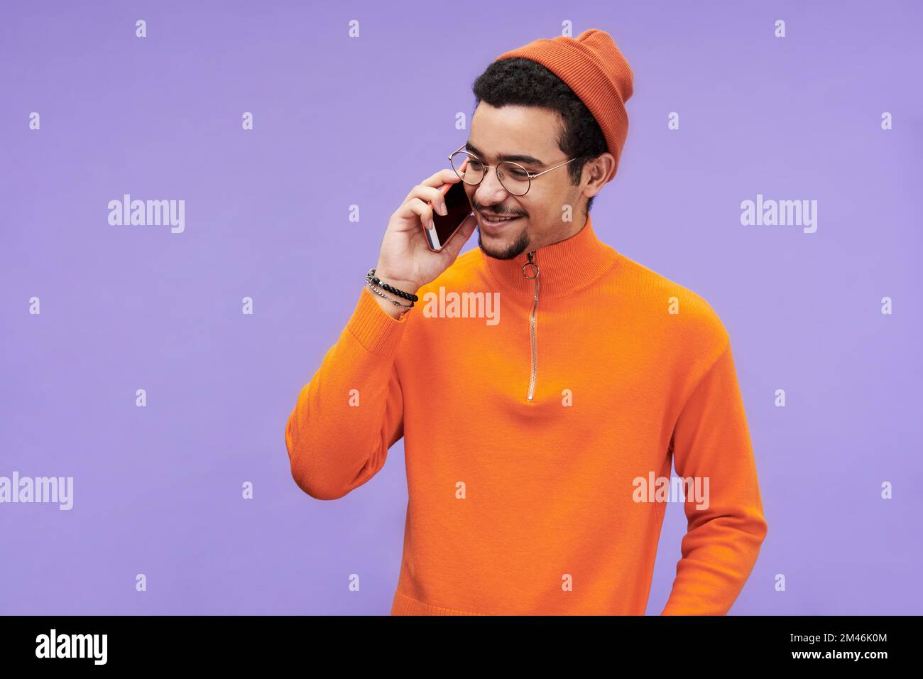 Joyeux gars en lunettes, pull-over orange et bonnet beanie parlant sur le téléphone mobile devant l'appareil photo sur fond violet Banque D'Images