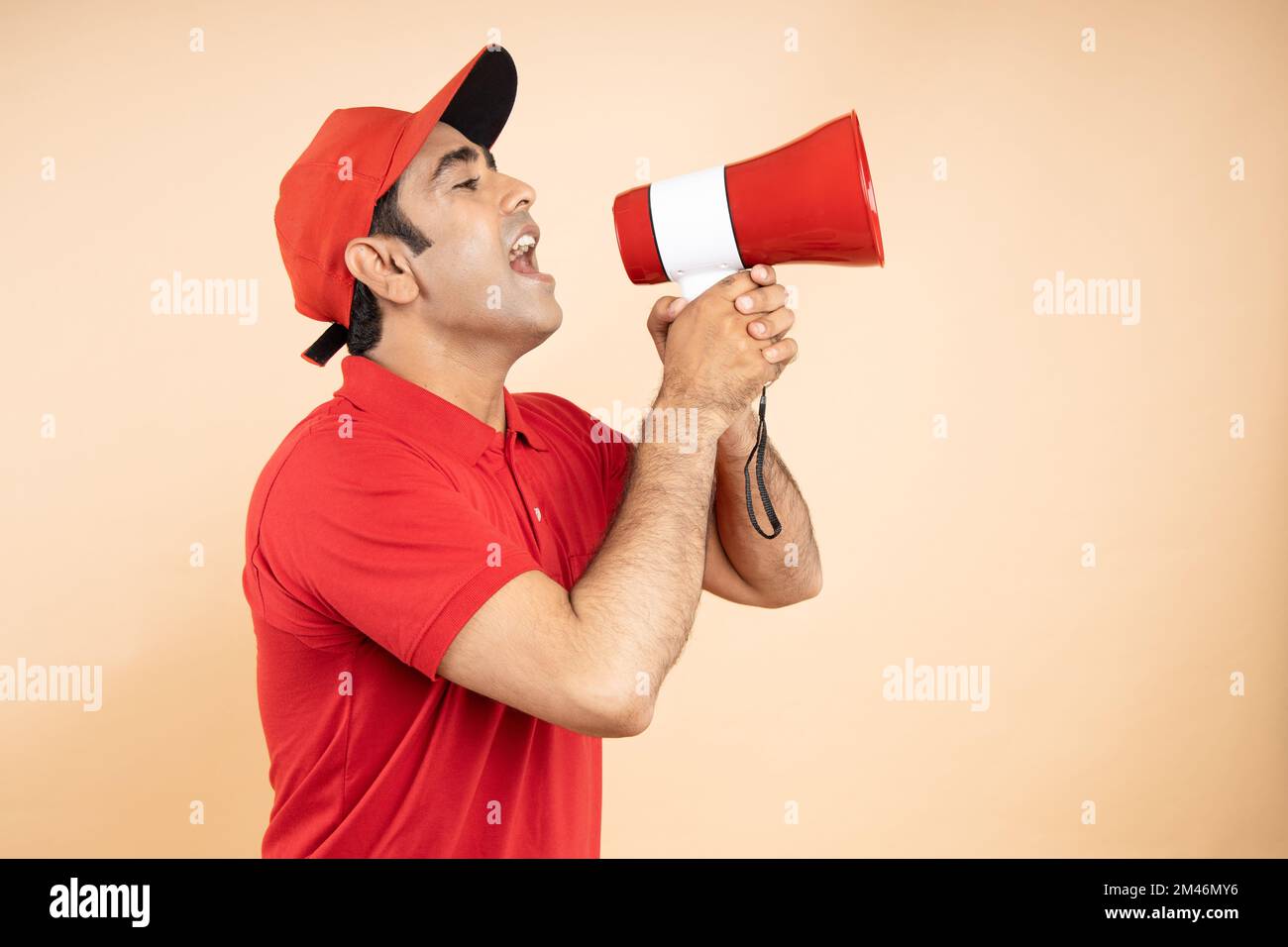 Heureux Indien excité livreur portant la casquette rouge et l'uniforme de T-shirt tenir criant dans le mégaphone ou haut-parleur isolé sur beige Studio backgroun Banque D'Images