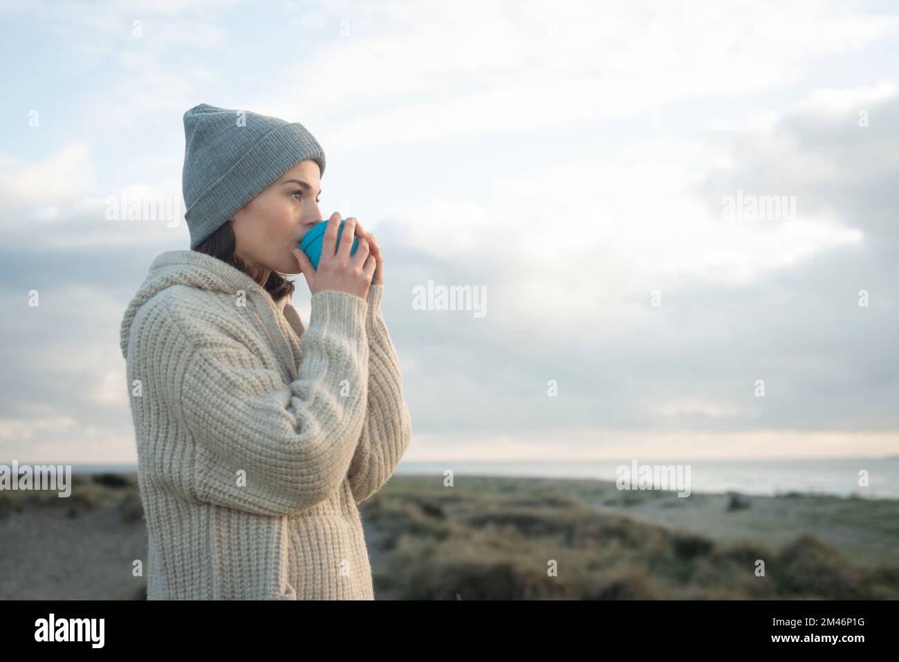 Femme à l'extérieur en tricots appréciant une boisson chaude tout en regardant la mer. Banque D'Images