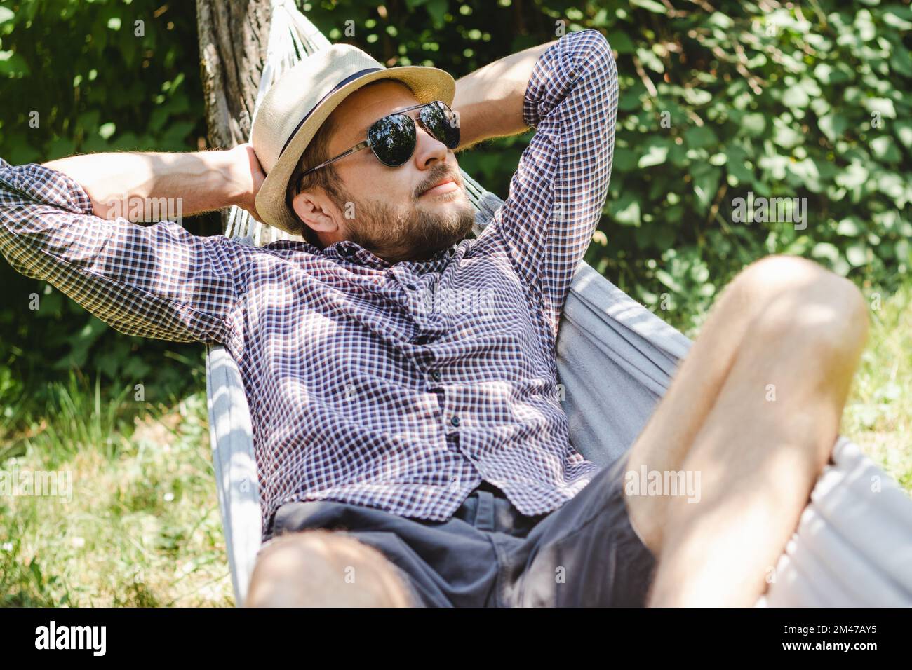 Homme barbu dans un chapeau et des lunettes de soleil couchés sur un hamac dans un jardin vert. Banque D'Images