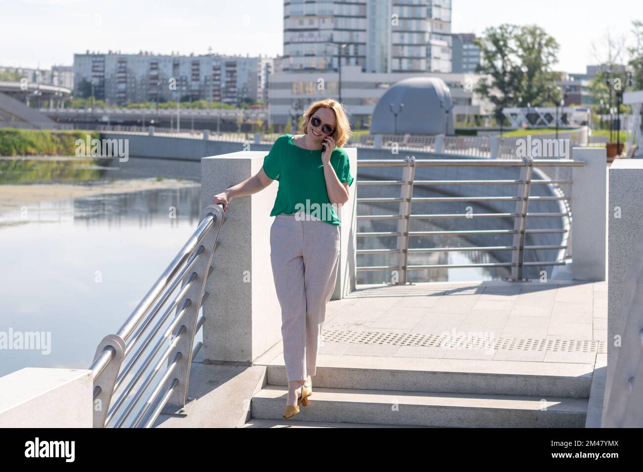 Portrait d'une fille étudiante souriante portant des lunettes de soleil, parlant sur un téléphone portable tout en marchant sur la rive. Formation à distance, formation en ligne a Banque D'Images