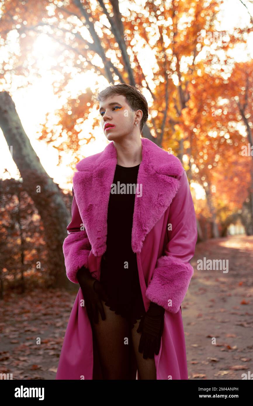 Auto-assuré jeune homme liquide de genre avec cheveux courts et maquillage dans le cuir fuchsia brillant et manteau de fourrure debout près des arbres d'automne et regardant sur Banque D'Images