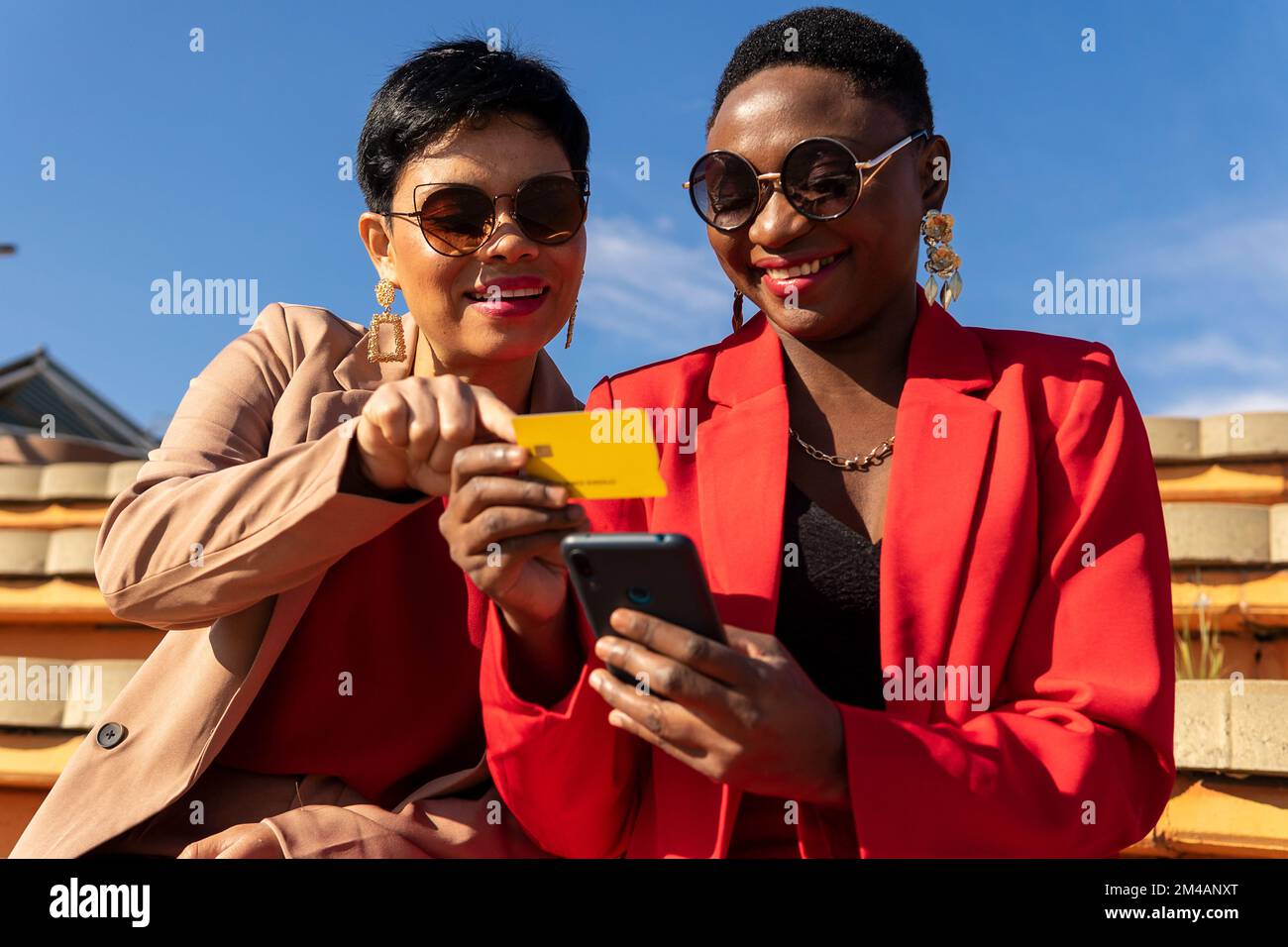 Positive femme hispanique adulte dans des vêtements élégants et des lunettes de soleil pointant à la carte de crédit tout en étant assis près de souriant ami afro-américain maki Banque D'Images