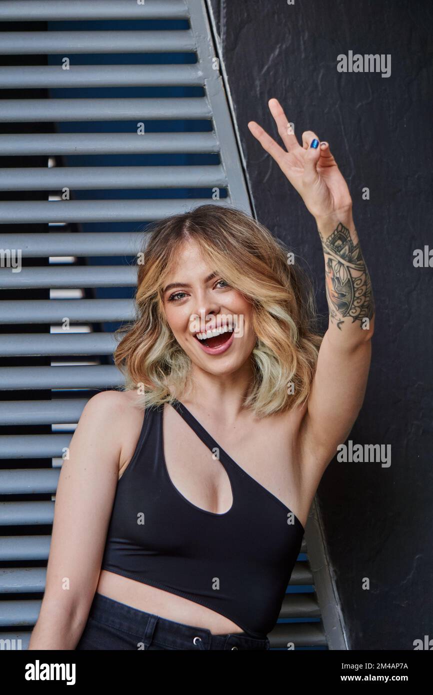 Jeune femme à l'assurance avec cheveux blonds ondulés et tatouage sur le bras dans un joli petit top souriant et montrant V geste dans la rue Banque D'Images