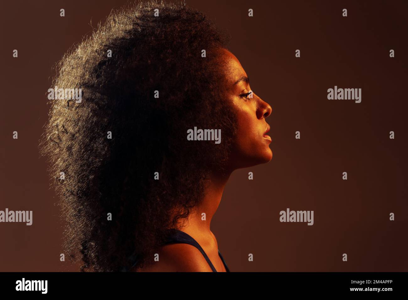 Vue latérale d'une femme afro-américaine pensive vêtu d'une robe colorée qui se tient debout dans un studio sombre, près d'un fond marron Banque D'Images