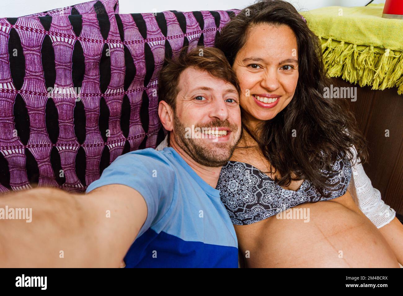 Portrait de selfie de couple latin, homme adulte caucasien avec barbe et yeux bleus avec sa femme, femme enceinte brésilienne adulte souriante, heureux assis sur sof Banque D'Images