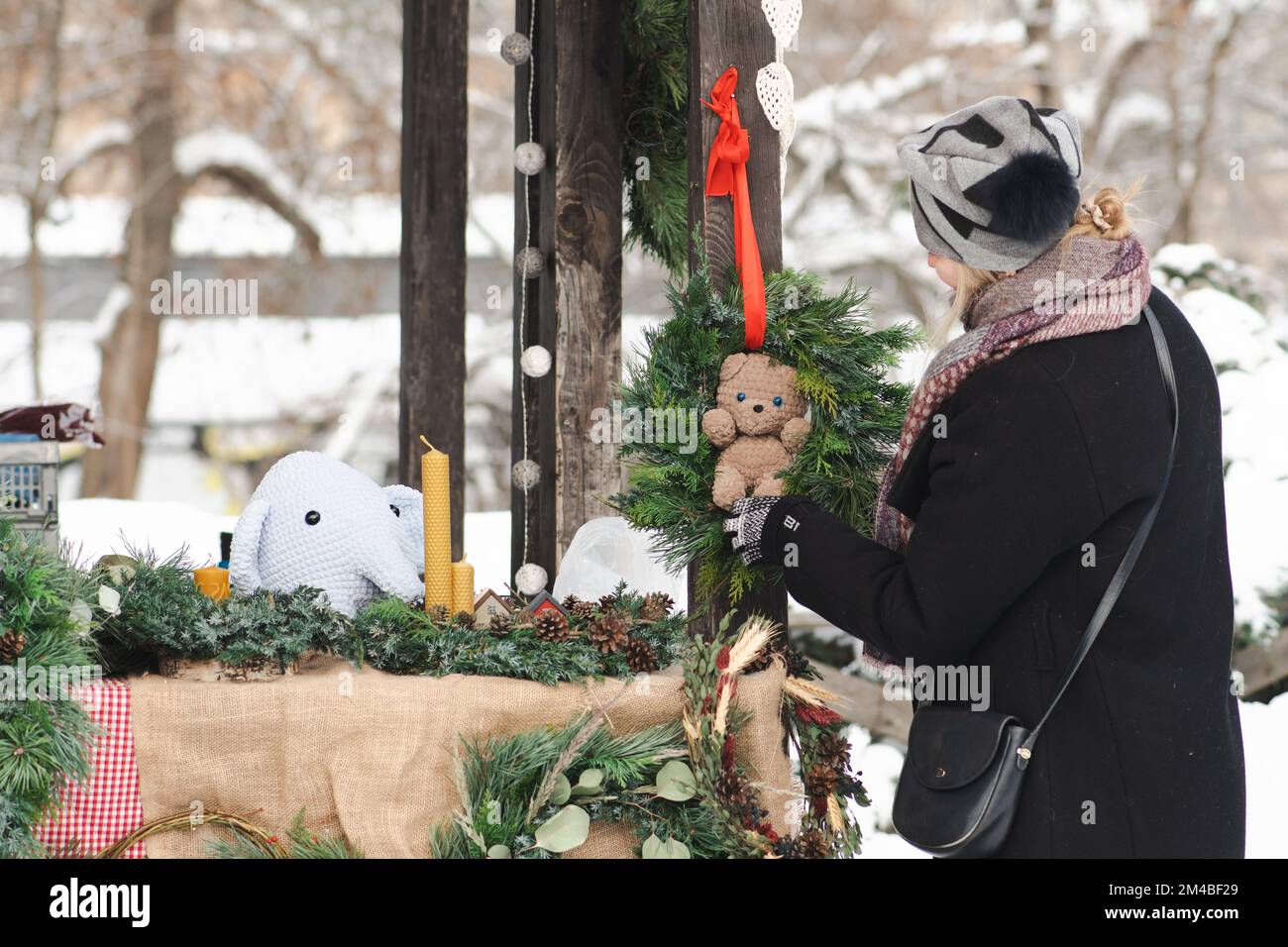 Belle fille en vêtements chauds achetant une guirlande de Noël ou une couronne avec ours en peluche dans un marché de rue en hiver Banque D'Images