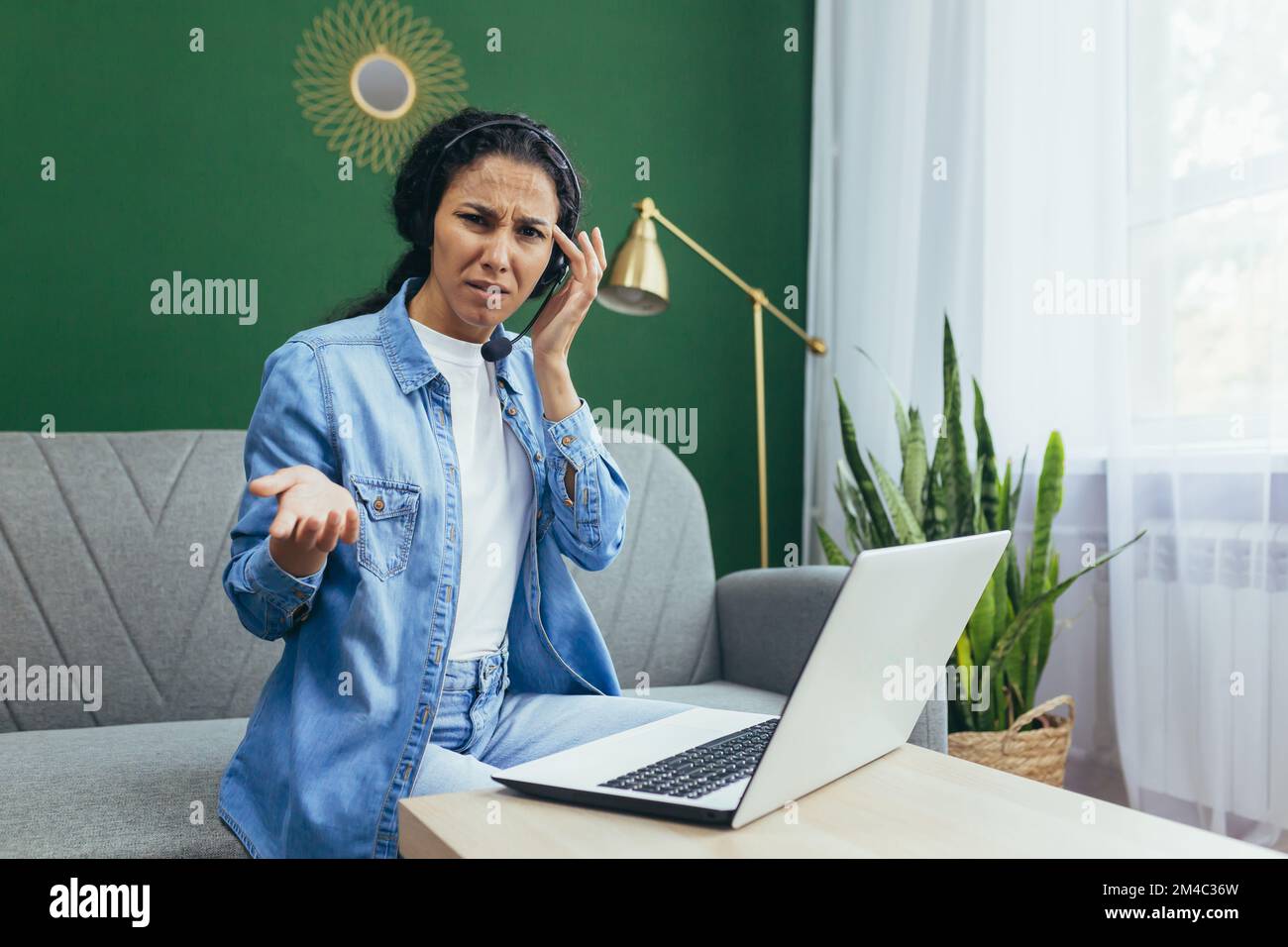 Portrait d'une femme frustrée et bouleversée à la maison, d'une femme hispanique travaillant à distance à regarder un ordinateur portable au travail et d'un casque d'appel vidéo assis sur un canapé dans le salon. Banque D'Images