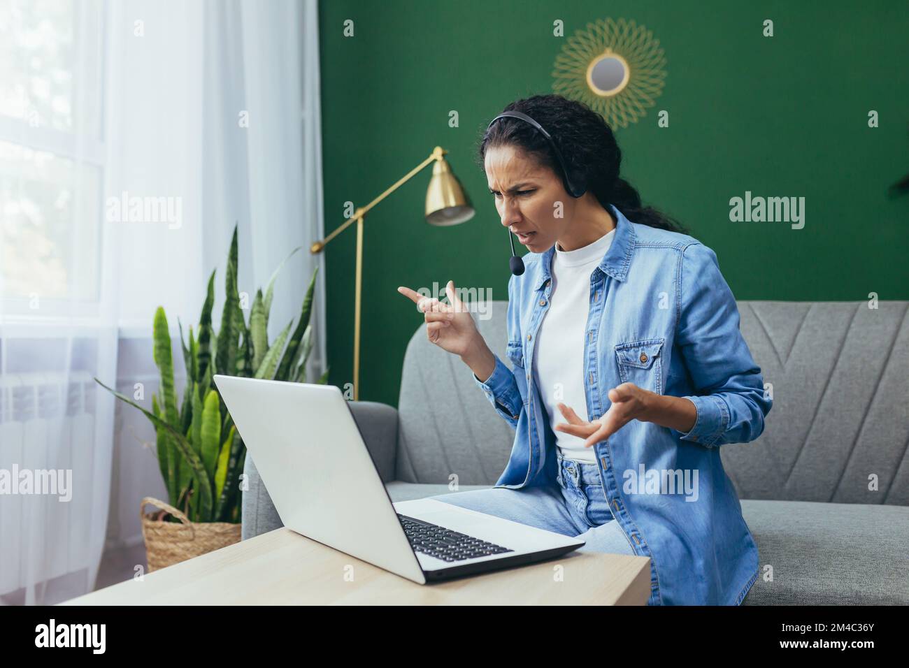 Portrait d'une femme frustrée et bouleversée à la maison, d'une femme hispanique travaillant à distance à regarder un ordinateur portable au travail et d'un casque d'appel vidéo assis sur un canapé dans le salon. Banque D'Images