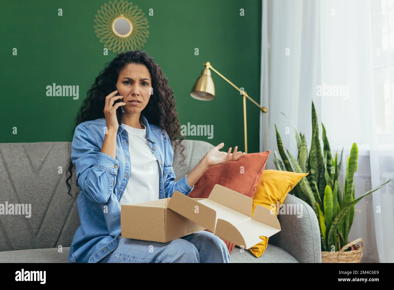 Une femme insatisfaite a reçu un colis erroné et endommagé, une femme hispanique regardant la caméra et parlant au service à la clientèle au téléphone, assis sur un canapé à la maison avec une boîte en carton. Banque D'Images