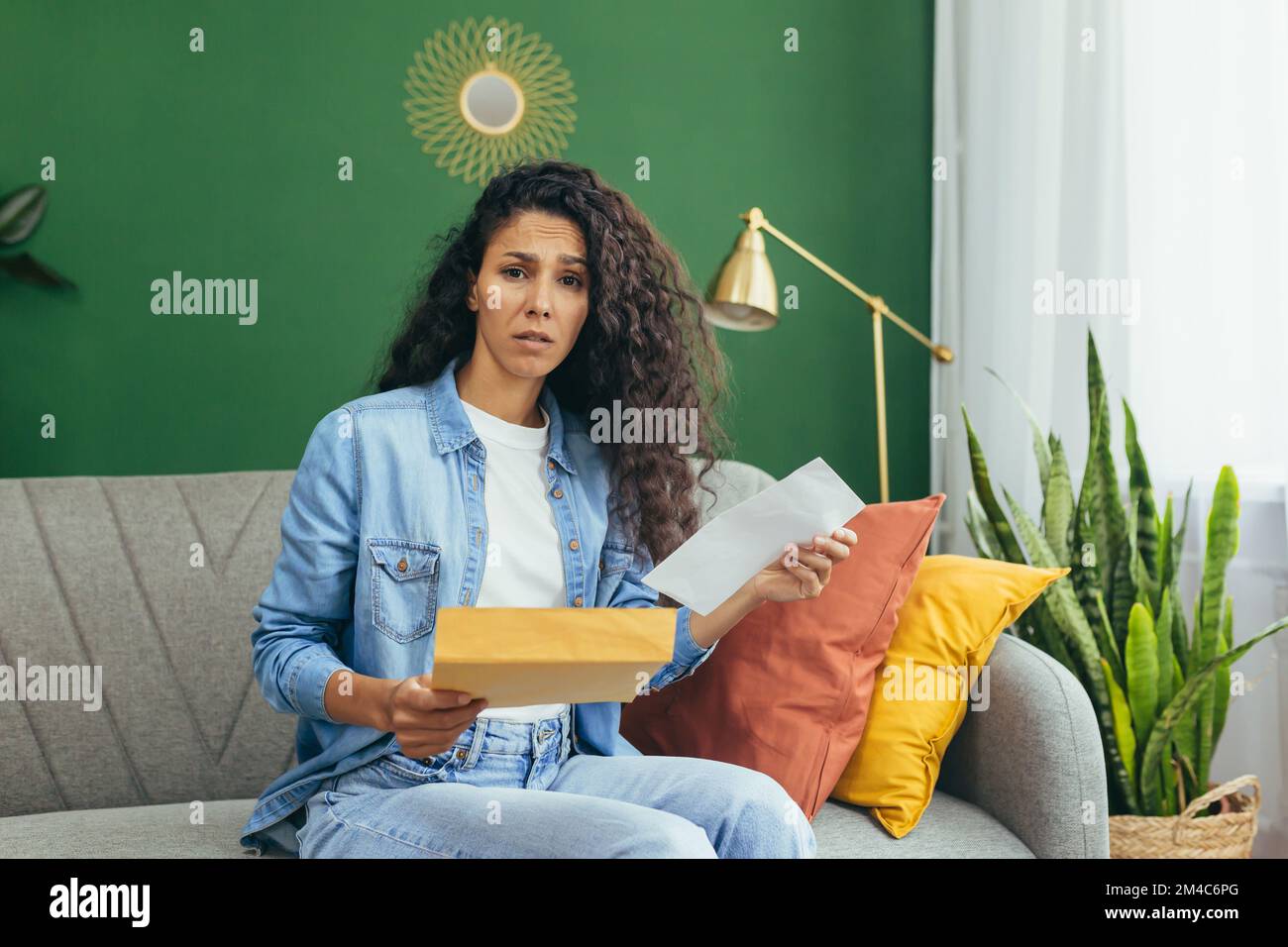 Portrait d'une femme déçue et triste, d'une femme hispanique avec une enveloppe reçue regardant passer du froid à l'appareil photo, assis sur un canapé dans le salon. Banque D'Images