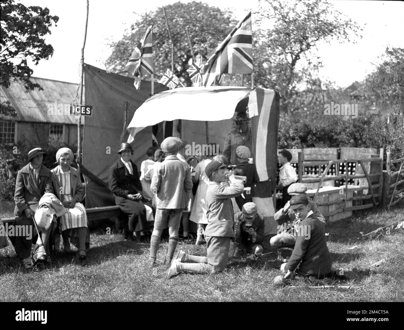 Vers la mi-1930s, historique, à une fête estivale dans la campagne, les mères et les écoliers, en shorts et en casquettes scolaires, se rassemblent dans un stand vendant des boissons non alcoolisées, Angleterre, Royaume-Uni. Les femmes portent les robes et les chapeaux de l'époque et plusieurs drapeaux de jack d'Union volent sur de longs poteaux, éventuellement élevés en célébration du couronnement des Britanniques, le roi George VI, qui a eu lieu en mai 1937. Banque D'Images