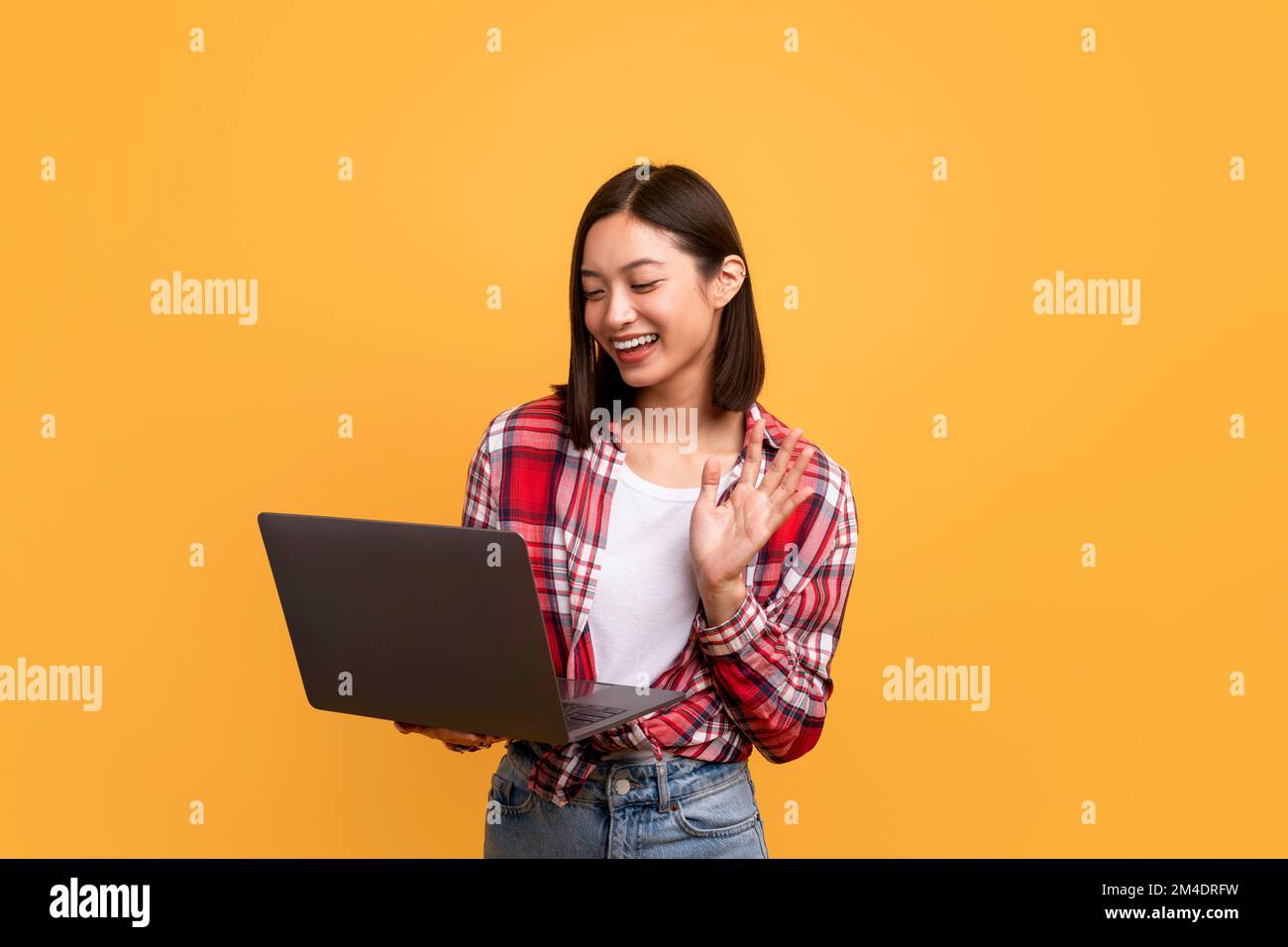 Appel vidéo en ligne. Bonne dame asiatique agitant à la webcam tenant et utilisant ordinateur portable sur fond jaune Banque D'Images