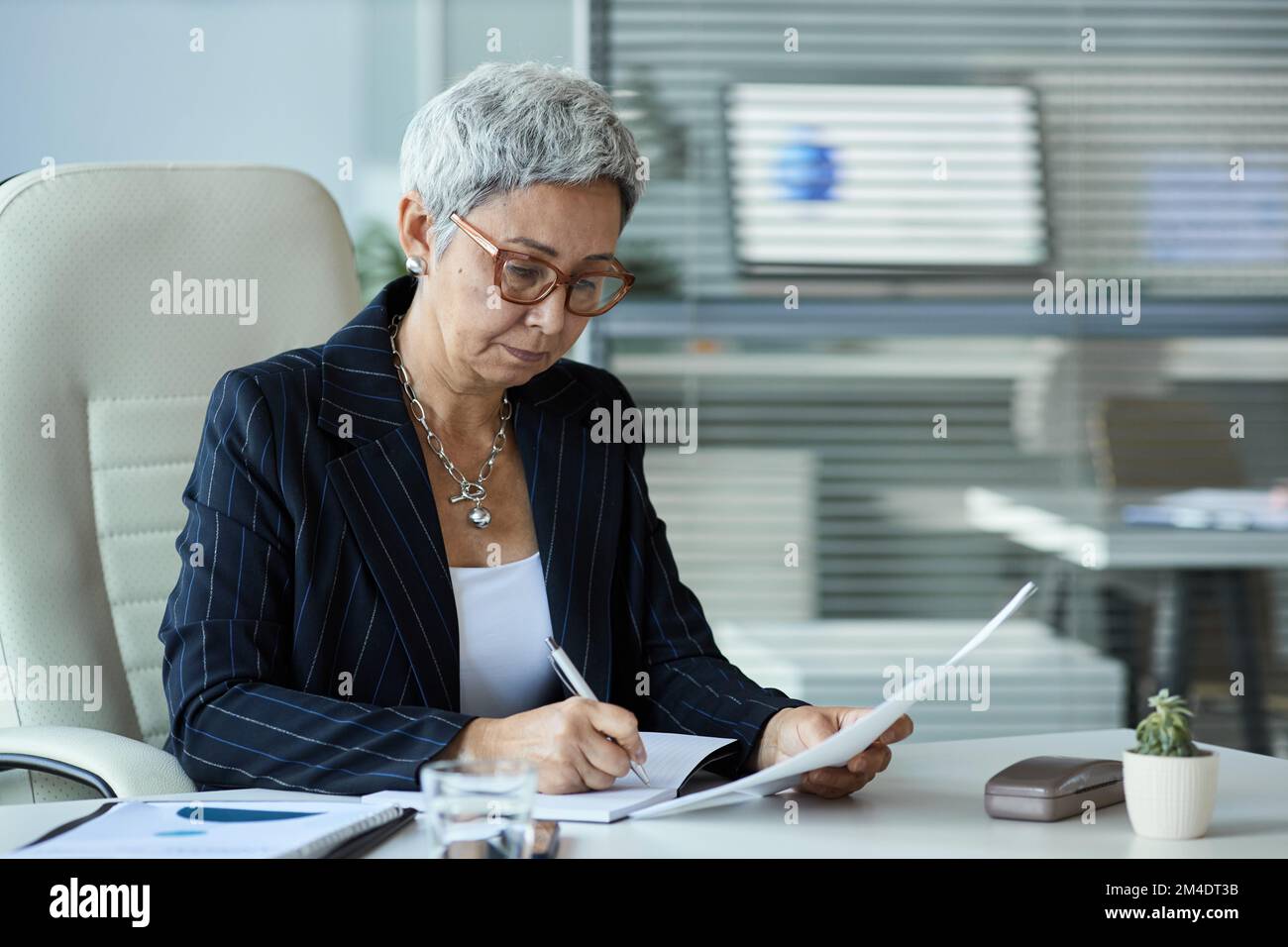 Portrait d'une femme âgée en tant que patron féminin assis au bureau et lisant un document portant des lunettes Banque D'Images