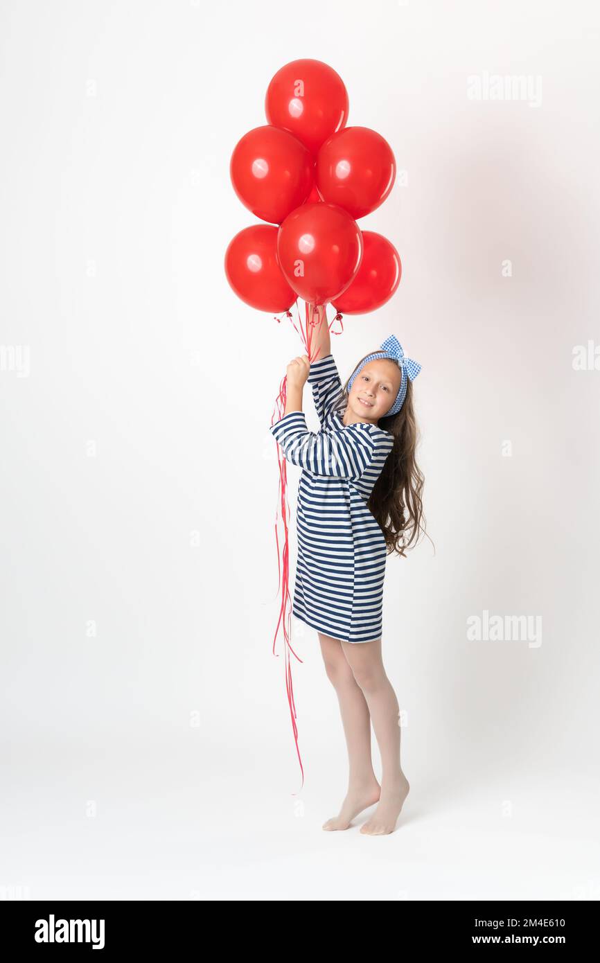 Fille tenant un grand bouquet de ballons rouges dans les mains, souriant et regardant l'appareil photo. Studio photo du modèle 10 ans en robe à rayures blanches et bleues Banque D'Images