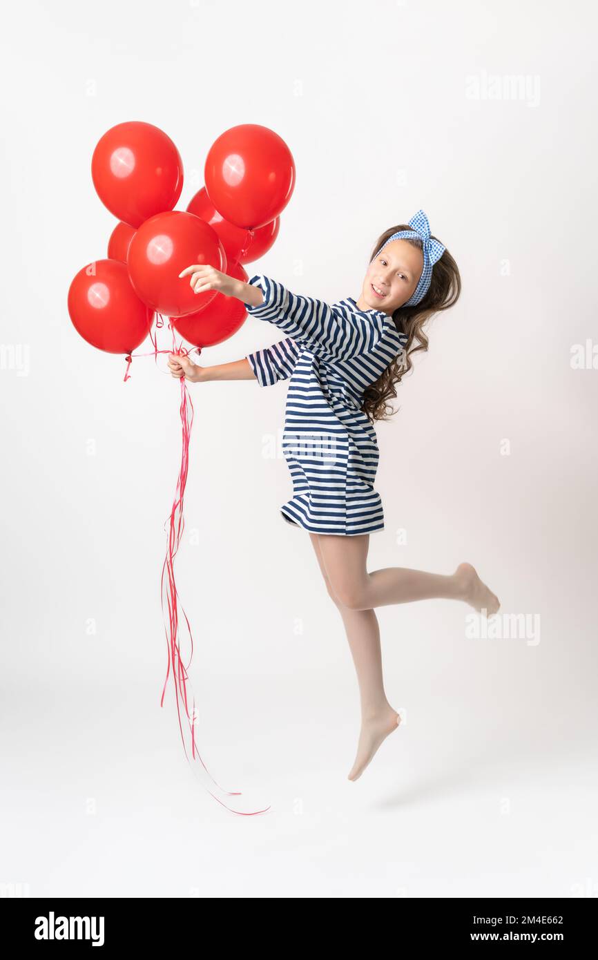 Une fille ludique en robe rayée saute, tenant un grand groupe de ballons rouges à la main et regardant l'appareil photo. Modèle féminin de dix ans avec cheveux longs Banque D'Images