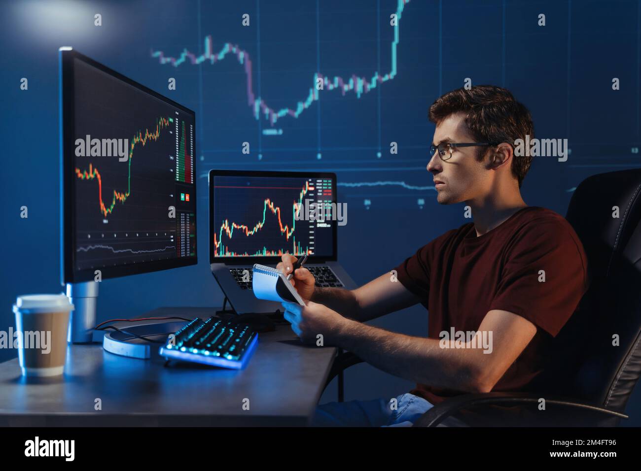 Vue latérale d'un courtier de crypto mâle assis sur son lieu de travail la nuit, mur numérique bleu avec arrière-plan du schéma financier. Commerçant vérifiant le chandelier ch Banque D'Images